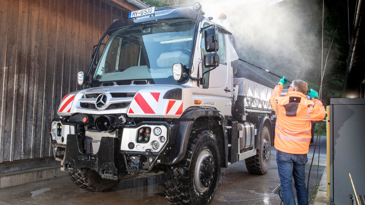 Čovek u narandžastoj radnoj jakni čisti srebrni Unimog uređajem za čišćenje pod pritiskom. Upravo prelazi preko prozora vozača. Iznad kamiona se vidi izmaglica od prskanja.