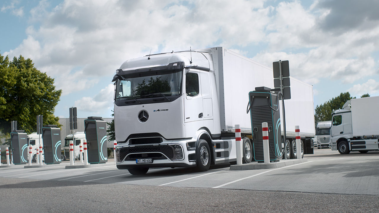 Beli eActros 600 sa ProCabin kompanije Mercedes‑Benz Trucks i belom prikolicom stoji na parkingu i puni se na stanici za punjenje. Oko kamiona vide se još nekoliko stanica za punjenje i saobraćajni stubići. U pozadini su još beli kamioni, a na levoj strani slike uočljivo je zeleno drveće pod delimično oblačnim nebom.