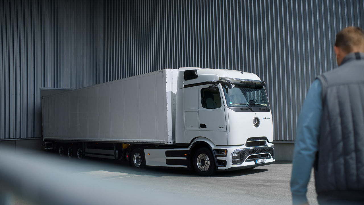 Beli eActros 600 sa ProCabinom kompanije Mercedes-Benz Trucks i dugom belom prikolicom stoji na asfaltiranom terenu ispred sivog zida od talasastog lima. Kamion se vidi sa strane, pri čemu je prednji deo blago okrenut ulevo. U prvom planu, okrenuta leđima prema posmatraču, stoji osoba u plavoj jakni i sivom prsluku koja gleda u kamion. Svetlo pada odozgo na scenu.