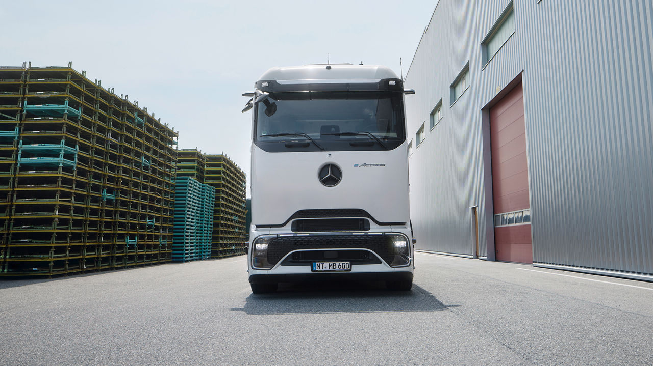 eActros 600 sa ProCabin kompanije Mercedes-Benz Trucks stoji frontalno na asfaltiranom terenu. Levo na slici vide se visoke gomile žutih i plavih paleta, dok se s desne strane nalazi velika, moderna zgrada sa sivom fasadom od talasastog lima i crvenim vratima. Nebo je svetlo i blago oblačno.
