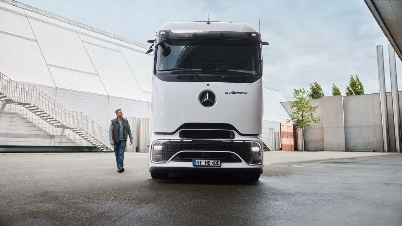 Muškarac u radnoj odeći hoda po asfaltiranoj površini, dok je u pozadini parkiran beli eActros 400 sa ProCabinom kompanije Mercedes-Benz Trucks. Levo se vidi moderna zgrada sa dugim spoljašnjim stepeništem, a desno drveće i druge građevinske strukture.