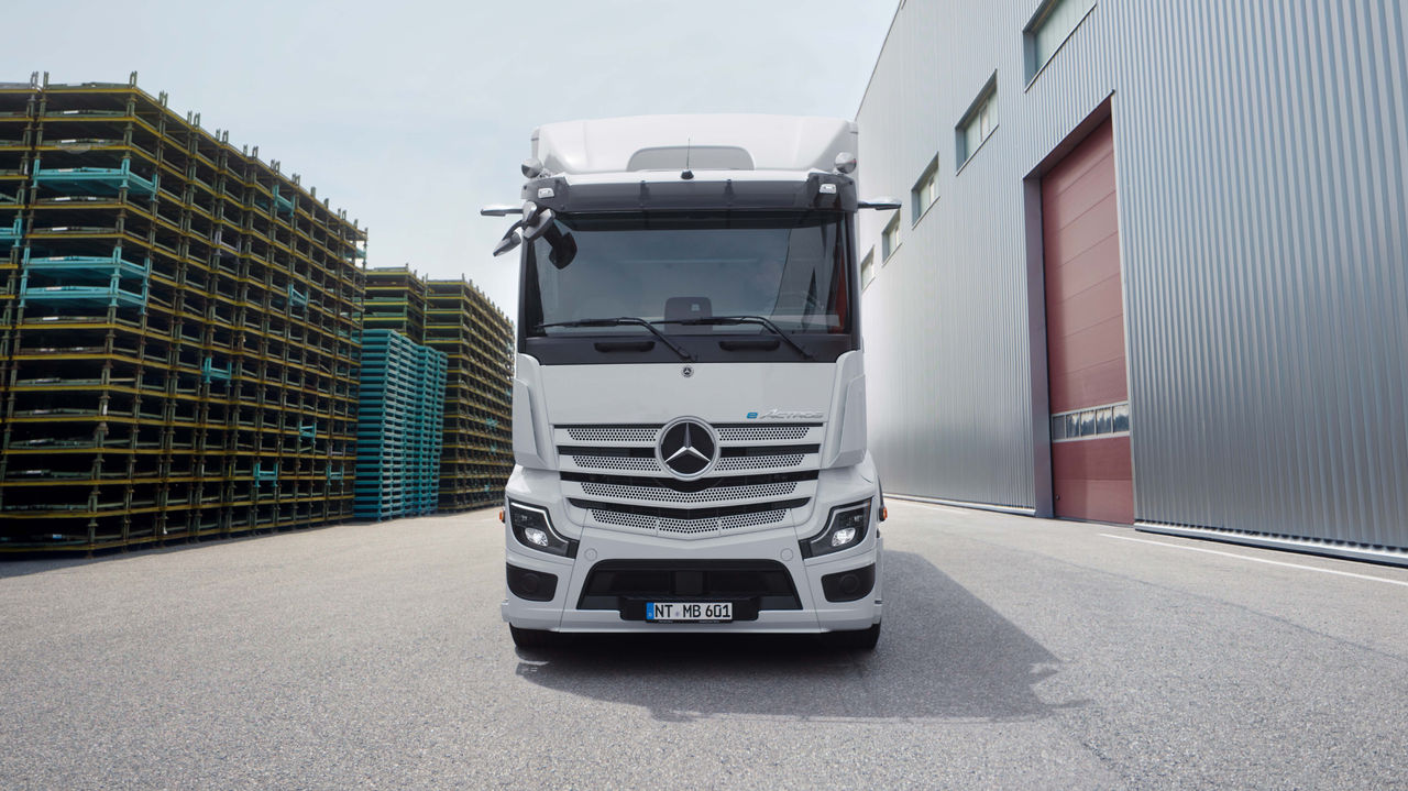 Jedan eActros 600 kompanije Mercedes-Benz Trucks stoji frontalno na asfaltiranoj površini ispred modernog objekta sa dugim spoljnim stepeništem. Kamion je beo i ima logo Mercedesove zvezde na maski hladnjaka, dok se u pozadini vide drveće i druge građevinske strukture.