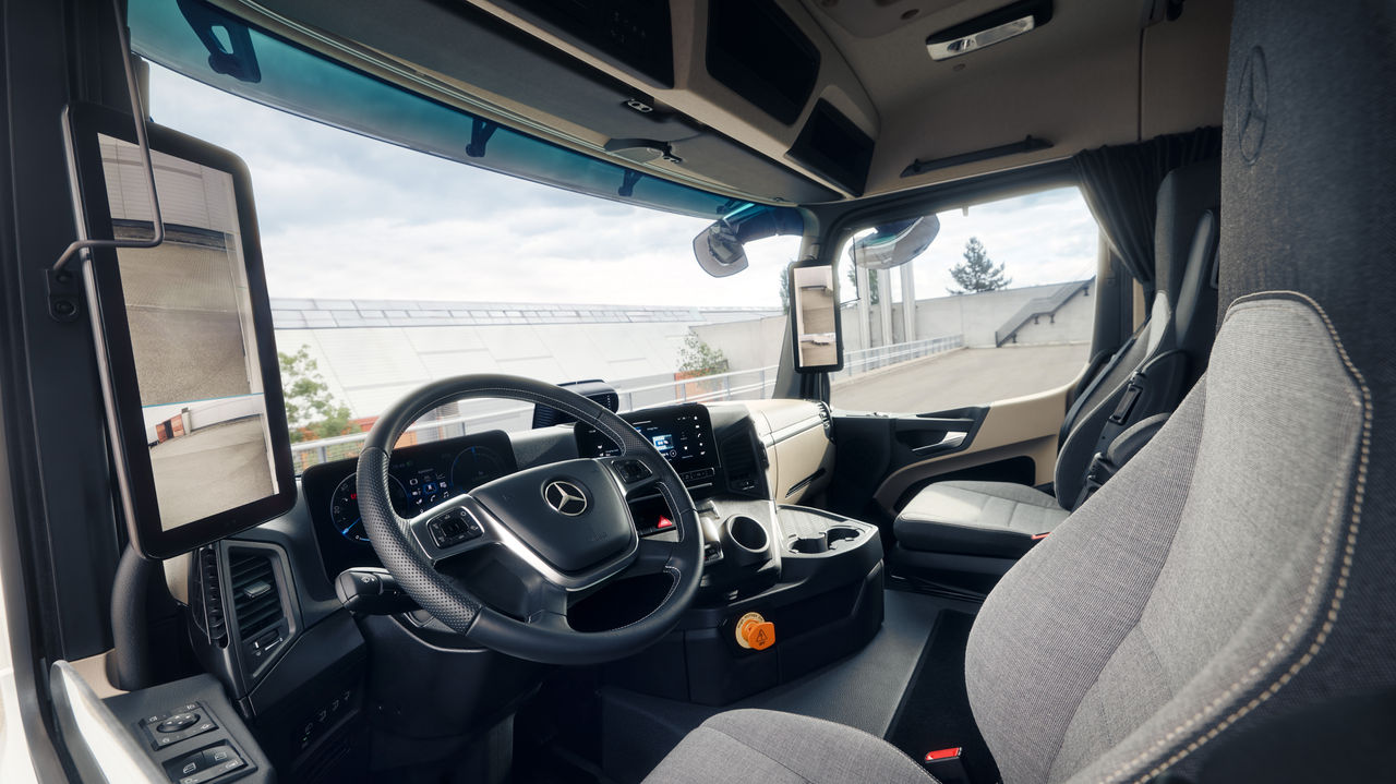 Slika prikazuje unutrašnjost eActros kompanije Mercedes-Benz Trucks iz perspektive suvozačevog sedišta. Vide se volan na kojem se nalazi Mercedes-Stern, instrument tabla sa digitalnim prikazima i ekranima, kao i udobna sedišta od svetlosive tkanine.