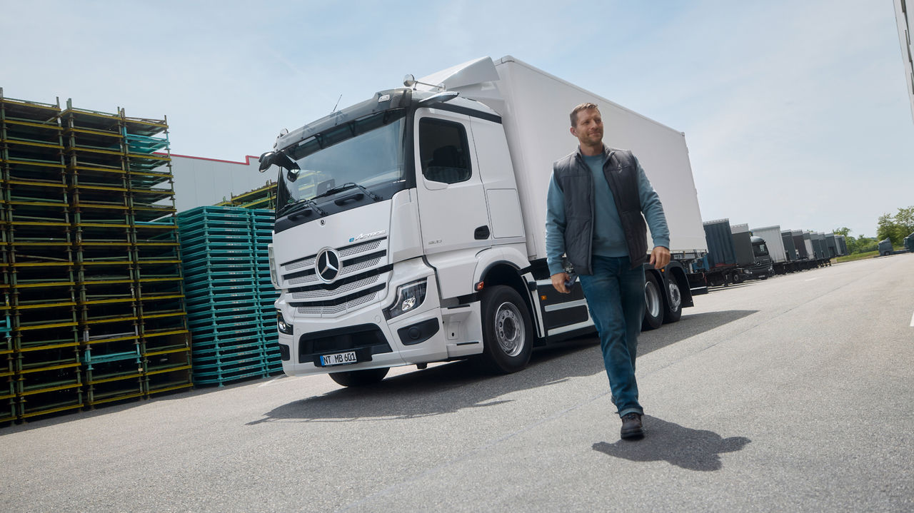 Muškarac u radnoj odeći hoda po asfaltiranoj površini, dok je u pozadini parkiran beli eActros 600 kompanije Mercedes-Benz Trucks sa prikolicom. Levo se vide složene palete, a desno velika zgrada koja ukazuje na skladišni ili industrijski kompleks.