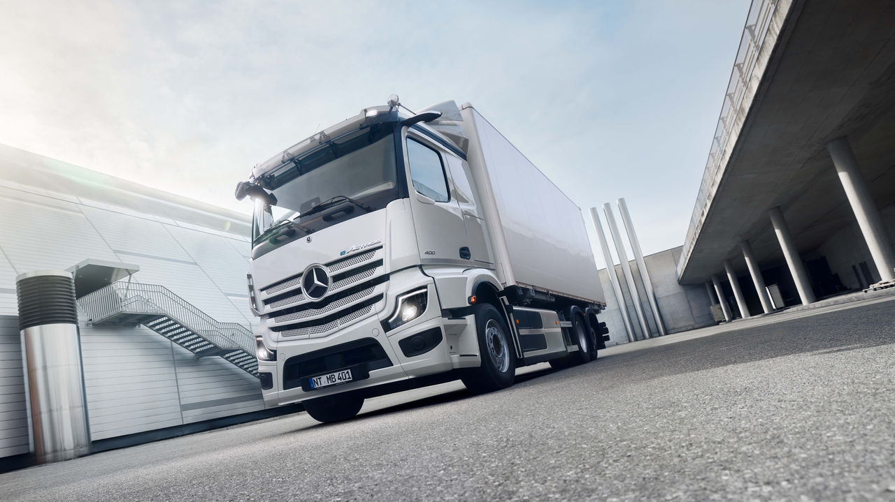 Na asfaltiranoj površini ispred modernog zdanja sa spoljnim stepeništem stoji beli eActros 400 marke Mercedes‑Benz Trucks, pri čemu sunce jako sija s leve strane odozgo. Desno na slici vidi se povišena betonska konstrukcija sa stubovima koja nagoveštava podvožnjak ili natkriveni prostor.