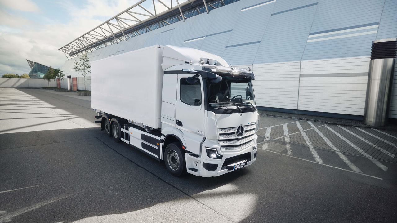 Beli eActros 400 kompanije Mercedes-Benz Trucks sa kutijastom nadogradnjom stoji na asfaltiranom parkiralištu ispred moderne zgrade sa staklenom fasadom. Sunčeva svetlost baca duge senke na tlo, što ukazuje na vedar dan.